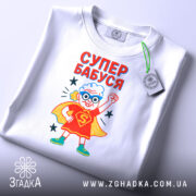 Футболка супер бабуся білого кольору, складена на столі, фон світло-сірий, принт з бабусею в червоному костюмі супергероя з синіми окулярами, текст 'СУПЕР БАБУСЯ' яскраво червоний, на футболці є етикетка з зеленим шнурком.