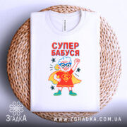 Футболка супер бабуся білого кольору, складена на плетеній кошику, фон білий, принт з бабусею в червоному костюмі супергероя з синіми окулярами, текст 'СУПЕР БАБУСЯ' яскраво червоний, зірочки навколо принта.