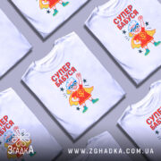 Футболка супер бабуся білого кольору, зображена на манекені, фон градієнтний сірий, принт з веселою бабусею в червоному костюмі супергероя з синіми окулярами та жовтим плащем, текст 'СУПЕР БАБУСЯ' яскраво червоний.