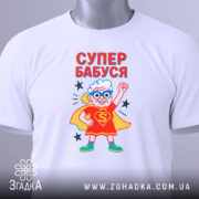 Футболка супер бабуся білого кольору, складена на столі, фон світло-сірий, принт з бабусею в червоному костюмі супергероя з синіми окулярами, текст 'СУПЕР БАБУСЯ' яскраво червоний, на футболці є етикетка з зеленим шнурком.
