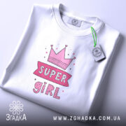 Футболка супер дівчина білого кольору, складена на столі, фон світло-сірий, принт з короною та написом 'SUPER GIRL' у рожевих тонах, з етикеткою на зеленому шнурку.
