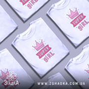 Футболка супер дівчина білого кольору, зображена з правого боку на манекені, фон світло-сірий, принт з короною та написом 'SUPER GIRL' у рожевих тонах.