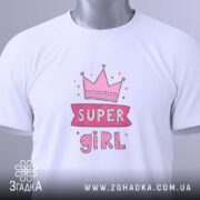 Футболка супер дівчина білого кольору, складена на столі, фон світло-сірий, принт з короною та написом 'SUPER GIRL' у рожевих тонах, з етикеткою на зеленому шнурку.