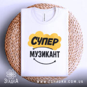 Футболка супер музикант, біла, складена на плетеній кошику, фон світло-сірий, принт жовтого кольору з написом "СУПЕР" та чорного з написом "МУЗИКАНТ", комірка видима.