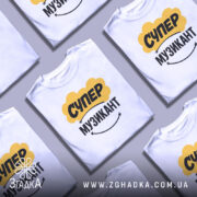 Футболка супер музикант, біла, зображена на манекені, фон світло-сірий, принт жовтого кольору з написом "СУПЕР" та чорного з написом "МУЗИКАНТ", під принтом є усміхнений смайлик.