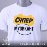 Футболка супер музикант, біла, складена на столі, фон світло-сірий, принт жовтого кольору з написом "СУПЕР" та чорного з написом "МУЗИКАНТ", на комірці є етикетка.