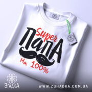 Футболка супер тато білого кольору, складена на поверхні, фон світло-сірий, принт з текстом 'Super ПАПА' червоного та чорного кольорів, з зеленим ярликом.