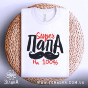 Футболка супер тато білого кольору, складена на плетеній кошику, фон світло-сірий, принт з текстом 'Super ПАПА' червоного та чорного кольорів.