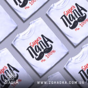 Футболка супер тато білого кольору, зображена з близького ракурсу на манекені, фон світло-сірий, принт містить текст 'Super ПАПА' червоного та чорного кольорів з малюнком вусів.