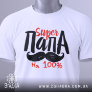 Футболка супер тато білого кольору, складена на поверхні, фон світло-сірий, принт з текстом 'Super ПАПА' червоного та чорного кольорів, з зеленим ярликом.