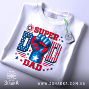 Футболка супер тато, біла, зображена зверху, складена на поверхні, фон світлий, принт яскравий з червоним, синім та білим кольорами, кулак з написом 'SUPER DAD' та елементами зірок.