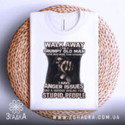 Футболка суворий старий, біла, зображена на плетеній кошику, фон світло-сірий, принт з чорним малюнком руки та текстом, що говорить про гнів і неприязнь до дурних людей.