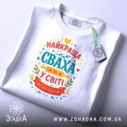 Футболка сваха білого кольору, складена на плоскій поверхні, з принтом на грудях, що містить текст "НАЙКРАЩА СВАХА У СВІТІ" в яскравих кольорах, на комірці видно етикетку.