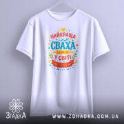 Футболка сваха білого кольору, підвішена на вішалці, з принтом на грудях, що містить текст "НАЙКРАЩА СВАХА У СВІТІ" в яскравих кольорах, фон світло-сірий.