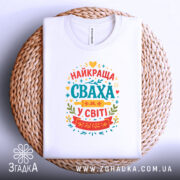 Футболка сваха білого кольору, складена на плетеній кошику, з принтом на грудях, що містить текст "НАЙКРАЩА СВАХА У СВІТІ" в яскравих кольорах, фон світлий.