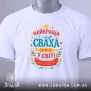 Футболка сваха білого кольору, складена на плоскій поверхні, з принтом на грудях, що містить текст "НАЙКРАЩА СВАХА У СВІТІ" в яскравих кольорах, на комірці видно етикетку.