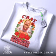 Футболка сват білого кольору, складена на столі, фон світло-сірий, принт з чоловіком у вишиванці, оточеним червоними трояндами та кольоровими квітами, напис 'СВАТ' зверху, на футболці є етикетка з зеленим шнурком.