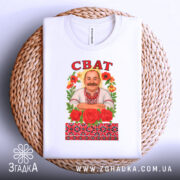 Футболка сват білого кольору, складена на плетеній кошику, фон світло-сірий, принт з чоловіком у вишиванці, оточеним червоними трояндами та кольоровими квітами, напис 'СВАТ' зверху.