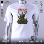Футболка свобода не дарма, біла, зображена на манекені, фон світло-сірий, принт з зображенням статуї Свободи та військових, текст 'FREEDOM IS NOT FREE' червоним кольором.