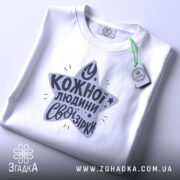 Футболка свої зірки. Складена біла футболка з круглим вирізом, зображена зверху. На футболці принт у формі зірки з текстом "У кожної людини свої зірки" у темно-сірому кольорі, з зеленим ярликом.