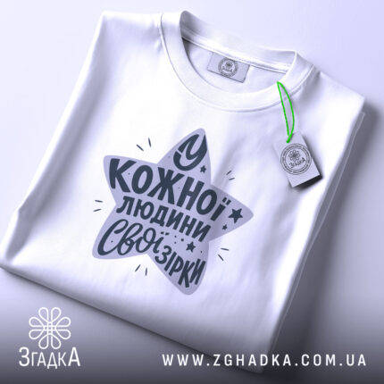 Футболка свої зірки. Складена біла футболка з круглим вирізом, зображена зверху. На футболці принт у формі зірки з текстом "У кожної людини свої зірки" у темно-сірому кольорі, з зеленим ярликом.
