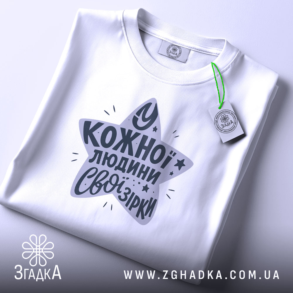 Футболка свої зірки. Складена біла футболка з круглим вирізом, зображена зверху. На футболці принт у формі зірки з текстом "У кожної людини свої зірки" у темно-сірому кольорі, з зеленим ярликом.