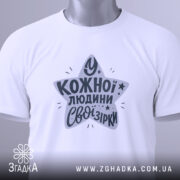 Футболка свої зірки. Складена біла футболка з круглим вирізом, зображена зверху. На футболці принт у формі зірки з текстом "У кожної людини свої зірки" у темно-сірому кольорі, з зеленим ярликом.