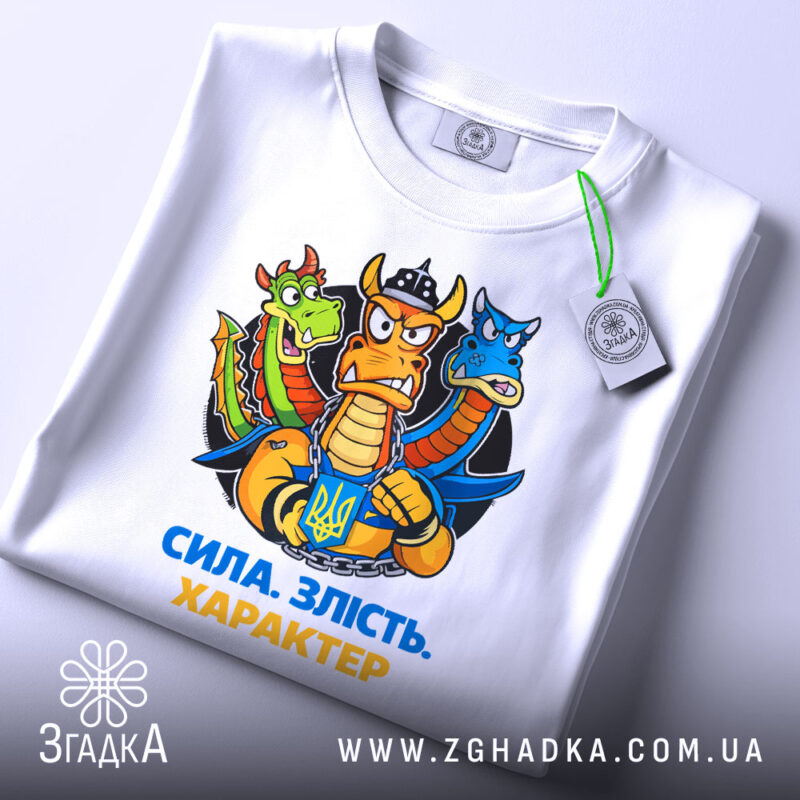 Футболка сила злість характер. Біла футболка, складена на поверхні, з принтом дракона, що тримає герб України. Принт яскравий, фон світло-сірий, на футболці є етикетка з зеленим шнурком.