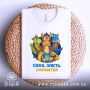 Футболка сила злість характер. Біла футболка, складена на плетеній кошику, з принтом дракона, що тримає герб України. Фон світло-сірий, деталі принта яскраві, акцент на емоційному виразі дракона.