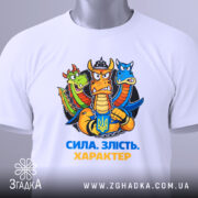 Футболка сила злість характер. Біла футболка, складена на поверхні, з принтом дракона, що тримає герб України. Принт яскравий, фон світло-сірий, на футболці є етикетка з зеленим шнурком.