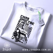 Футболка сила зростає в мені, біла, складена на поверхні, фон світлий, принт з чоловіком у спортивному костюмі з гантелями, чорний і білий кольори, текст "СИЛА ЗРОСТАЄ В МЕНІ", на ярлику зелена стрічка.