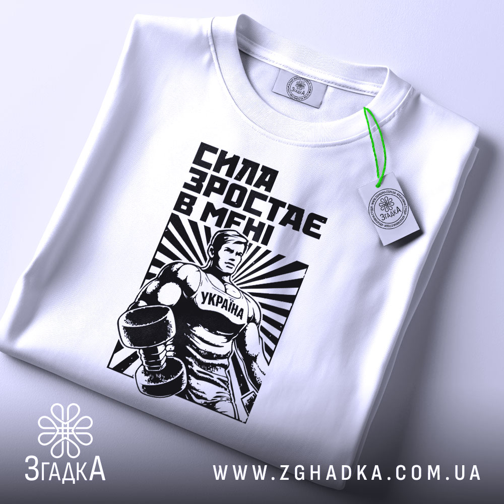 Футболка сила зростає в мені, біла, складена на поверхні, фон світлий, принт з чоловіком у спортивному костюмі з гантелями, чорний і білий кольори, текст "СИЛА ЗРОСТАЄ В МЕНІ", на ярлику зелена стрічка.