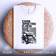 Футболка сила зростає в мені, біла, складена на плетеній кошику, фон світлий, принт з чоловіком у спортивному костюмі з гантелями, чорний і білий кольори, текст "СИЛА ЗРОСТАЄ В МЕНІ".