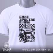 Футболка сила зростає в мені, біла, складена на поверхні, фон світлий, принт з чоловіком у спортивному костюмі з гантелями, чорний і білий кольори, текст "СИЛА ЗРОСТАЄ В МЕНІ", на ярлику зелена стрічка.