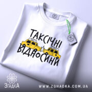 Футболка таксічні відносини, білого кольору, складена на столі, фон світло-сірий, принт з жовтими таксі та чорним текстом, з зеленим ярликом.