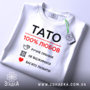 Футболка тато білого кольору, зображена зверху, складена на плоскій поверхні, фон світло-сірий, принт чорного та червоного кольорів з написом 'TATO 100% ЛЮБОВ' та символами догляду за одягом, з зеленим ярликом.