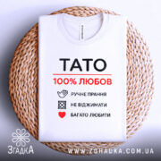 Футболка тато білого кольору, зображена зверху, складена на плетеній тарілці, фон світло-сірий, принт чорного та червоного кольорів з написом 'TATO 100% ЛЮБОВ' та символами догляду за одягом.