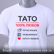 Футболка тато білого кольору, зображена зверху, складена на плоскій поверхні, фон світло-сірий, принт чорного та червоного кольорів з написом 'TATO 100% ЛЮБОВ' та символами догляду за одягом, з зеленим ярликом.