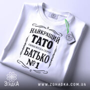 Футболка тато білого кольору, складена на поверхні, фон світло-сірий, принт чорного кольору з написом 'НАЙКРАЩИЙ ТАТО ОБМЕЖЕНА СЕРІЯ БАТЬКО №1', з зеленим ярликом.