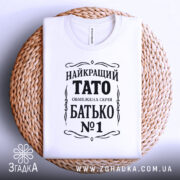Футболка тато білого кольору, складена на плетеній кошику, фон світло-сірий, принт чорного кольору з написом 'НАЙКРАЩИЙ ТАТО ОБМЕЖЕНА СЕРІЯ БАТЬКО №1'.