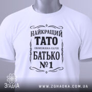 Футболка тато білого кольору, складена на поверхні, фон світло-сірий, принт чорного кольору з написом 'НАЙКРАЩИЙ ТАТО ОБМЕЖЕНА СЕРІЯ БАТЬКО №1', з зеленим ярликом.