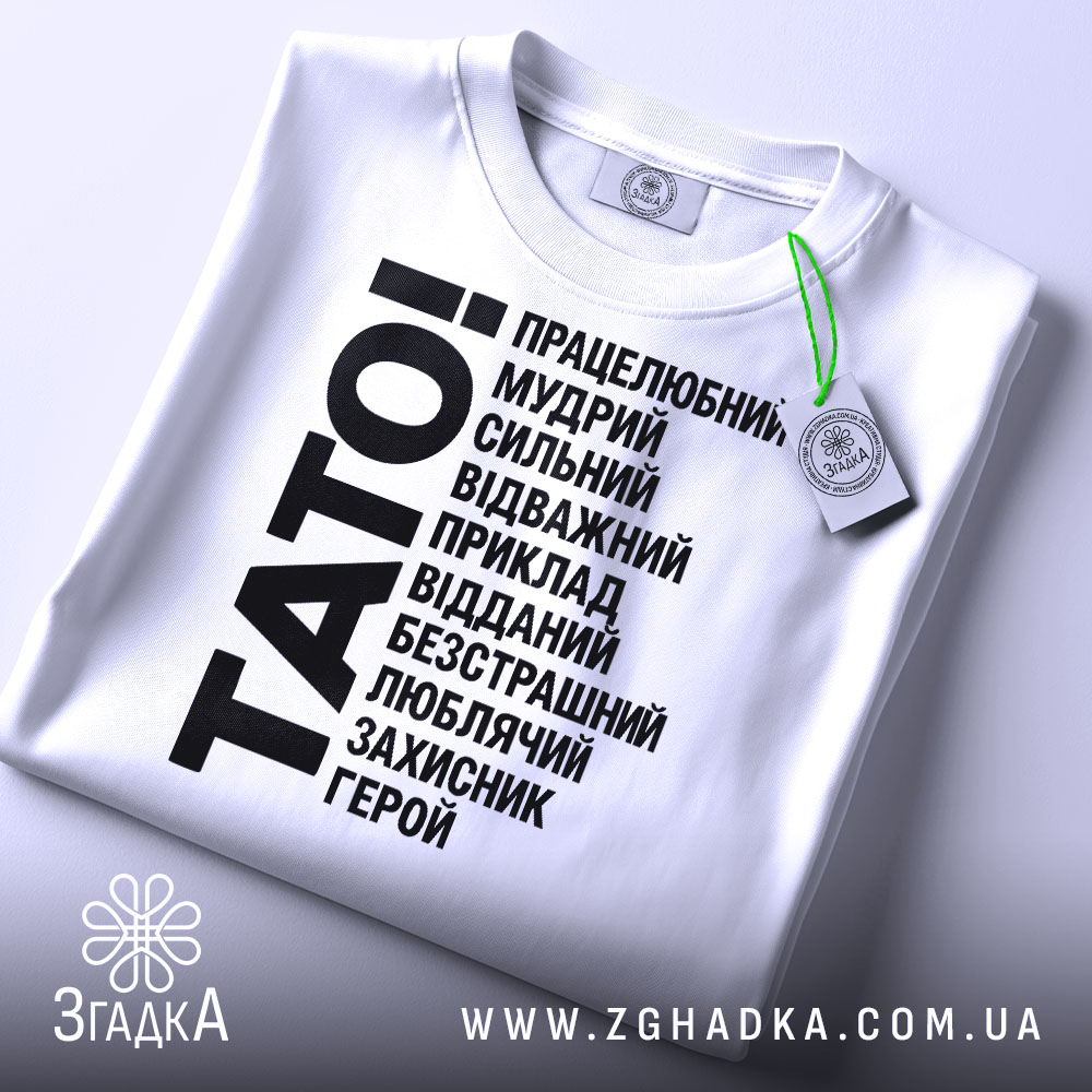 Футболка тато білого кольору, складена на поверхні, фон світло-сірий, принт чорного кольору з написом 'TATO!' та переліком позитивних якостей тата, на комірці є етикетка.