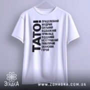 Футболка тато білого кольору, підвішена на вішалці, фон світло-сірий, принт чорного кольору з написом 'TATO!' та переліком позитивних якостей тата.