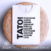 Футболка тато білого кольору, складена на плетеній кошику, фон світлий, принт чорного кольору з написом 'TATO!' та переліком позитивних якостей тата.