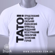Футболка тато білого кольору, складена на поверхні, фон світло-сірий, принт чорного кольору з написом 'TATO!' та переліком позитивних якостей тата, на комірці є етикетка.
