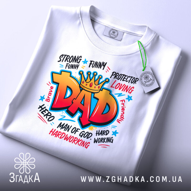 Футболка тато супергерой білого кольору, складена на поверхні, фон світло-сірий, принт яскравий з написом 'DAD' в червоному кольорі, з зеленим ярликом, що звисає з коміра.