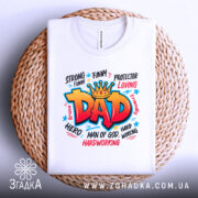 Футболка тато супергерой білого кольору, складена на плетеній кошику, фон світло-сірий, принт яскравий з написом 'DAD' в червоному кольорі, оточений словами, що описують позитивні якості тата.