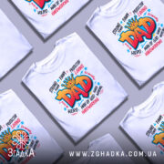 Футболка тато супергерой білого кольору, зображена на манекені, фон світло-сірий, принт яскравий з написом 'DAD' в червоному кольорі, оточений словами, що описують позитивні якості тата.