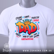 Футболка тато супергерой білого кольору, складена на поверхні, фон світло-сірий, принт яскравий з написом 'DAD' в червоному кольорі, з зеленим ярликом, що звисає з коміра.