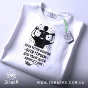 Футболка тато білого кольору, складена на поверхні, фон світло-сірий, принт чорного кольору з написом 'БУТИ ТАТОМ ОЗНАЧАЄ БУТИ ГЕРОЄМ ДЛЯ СВОЇХ ДІТЕЙ КОЖНОГО ДНЯ' та зображенням батька з дитиною на плечах, з зеленим ярликом.